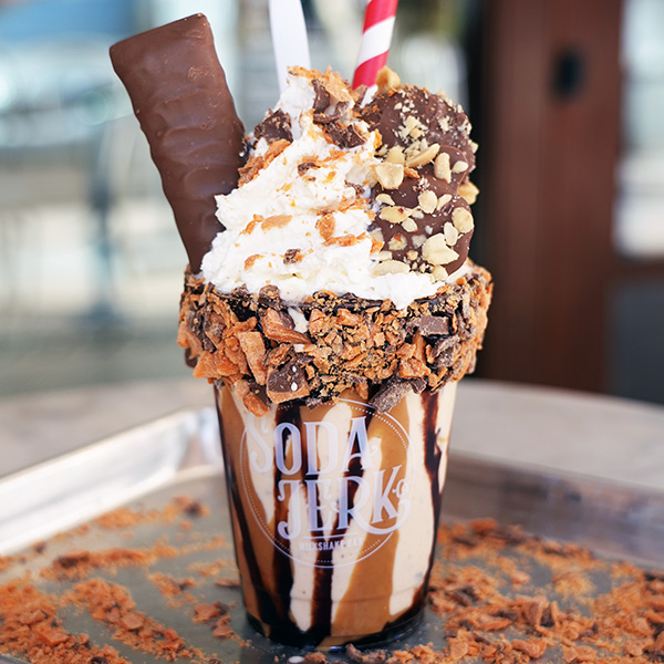 Butter Fingers Killer Shake in a Soda Jerk cup surrounded by Butterfinger crumbs.